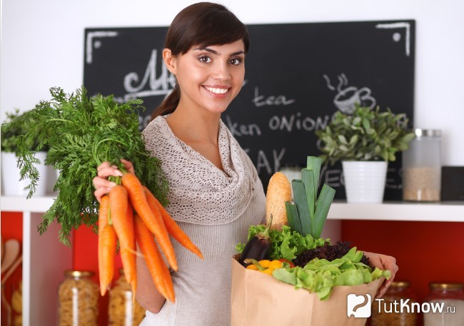 Девушка разбирает пакет с продуктами и держит морковь